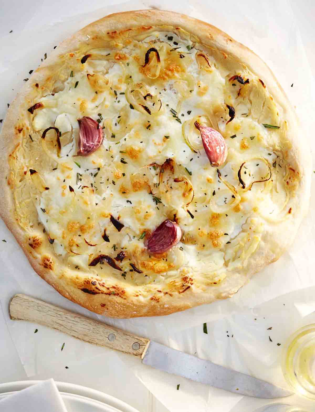 A rosemary red onion pizza with ricotta cheese, garlic cloves, and Parmesan cheese on parchment paper.