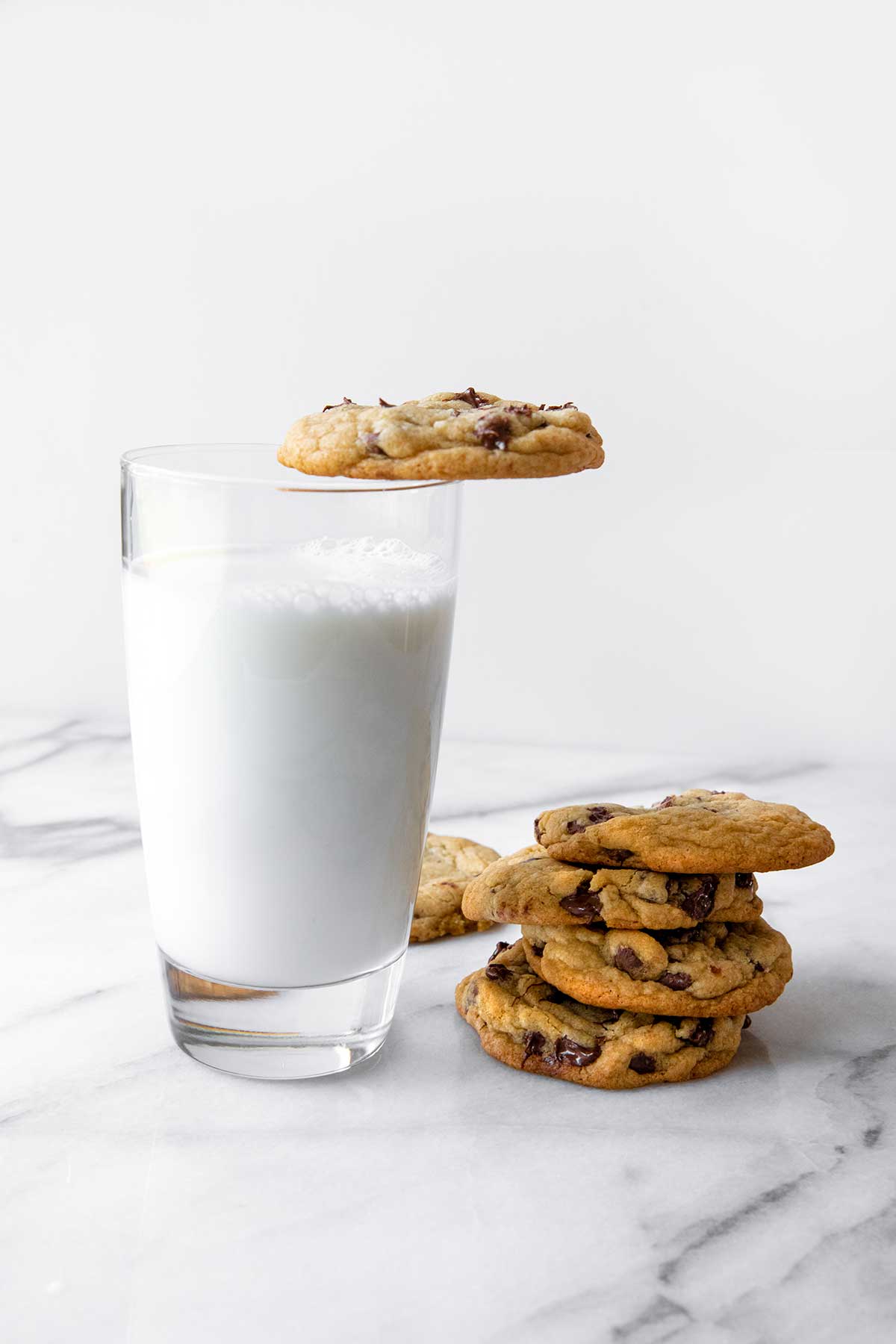 A glass of milk with a chocolate chip cookie on top and several others next to it.