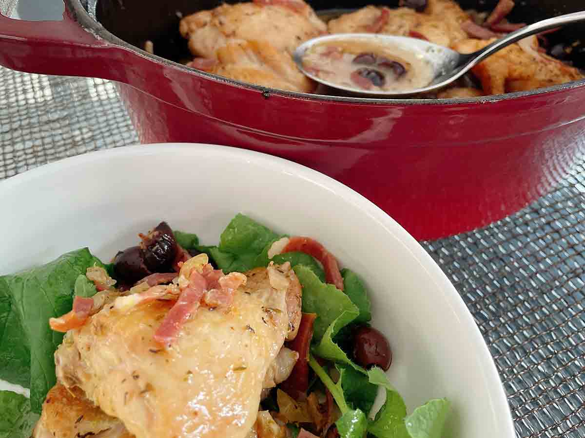 A white bowl with a braised chicken thighs on top of greens, and a red Dutch oven in the background containing more chicken.