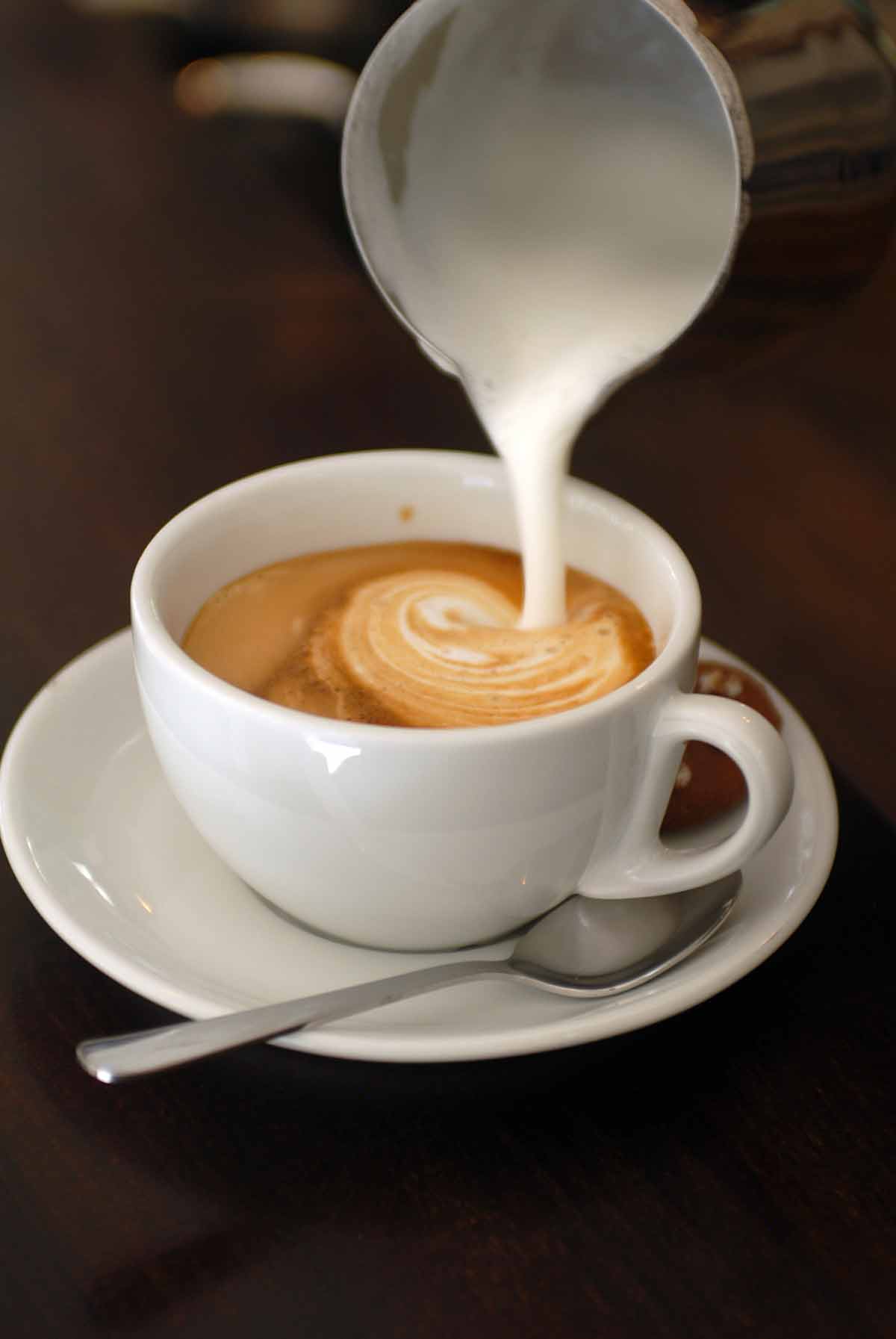 A cup of cappuccino on a saucer with milk being poured into it.