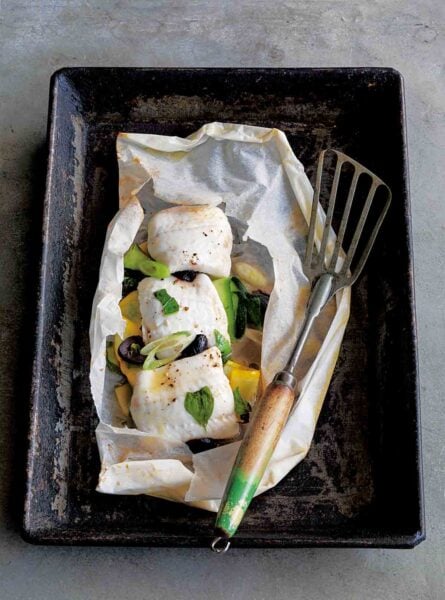 A rimmed baking sheet with fish in parchment and a fish spatula resting beside it.