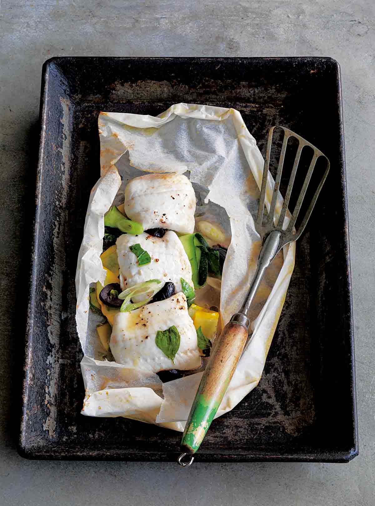 A rimmed baking sheet with fish in parchment and a fish spatula resting beside it.