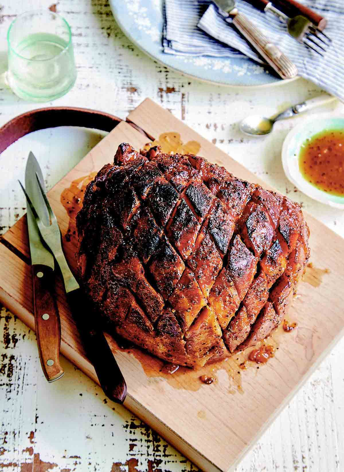 A cooked ham with broiled crispy skin on a wooden cutting board with a dish of sauce on the side.