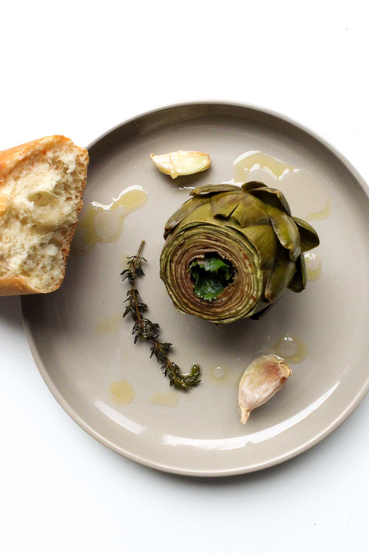 A braised artichoke on a round plate with a couple of garlic cloves, a thyme sprig, and piece of bread on the side.