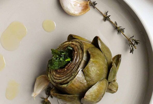 Braised artichokes on round plates with garlic and thyme sprigs and torn chunks of bread on the side.