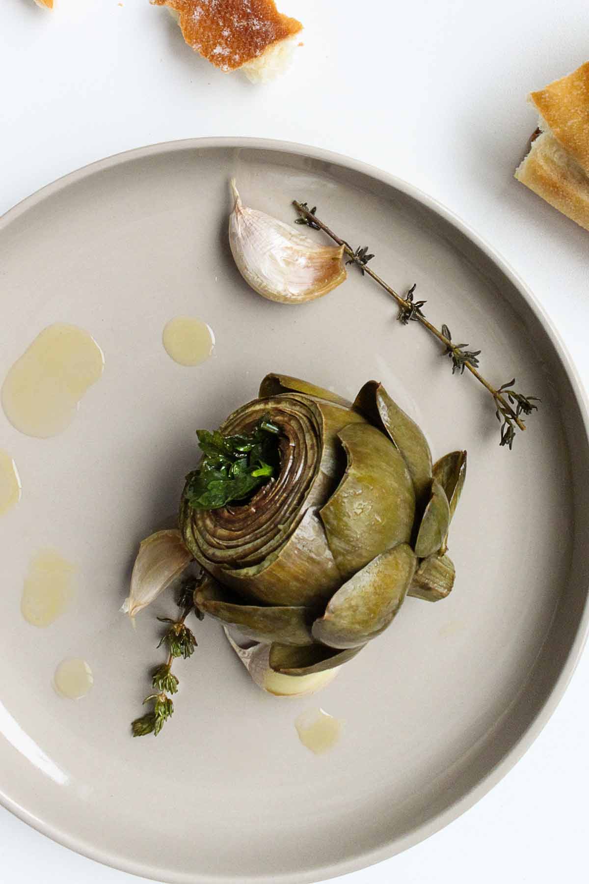 Braised artichokes on round plates with garlic and thyme sprigs and torn chunks of bread on the side.