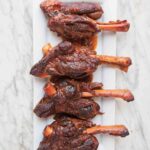 Four lamb shanks with coffee and ancho chile on a rectangular white plate on a white and grey marble surface.