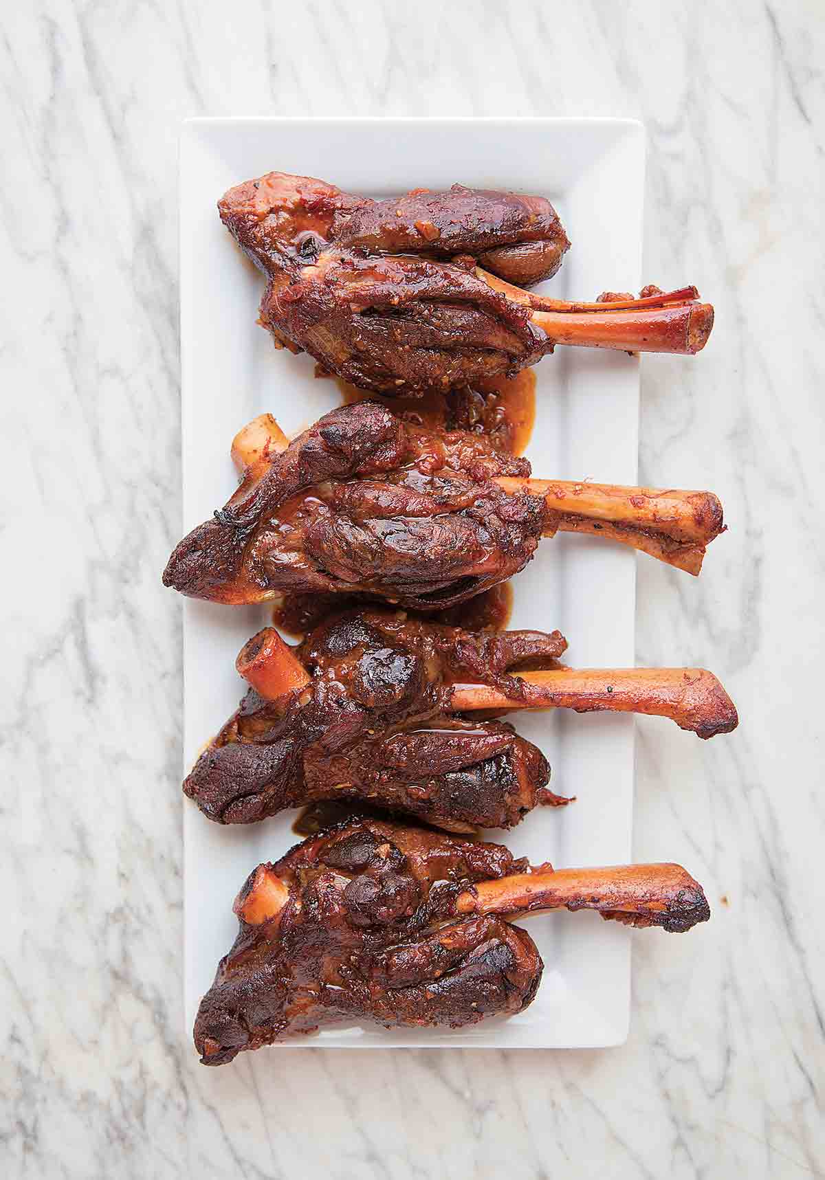 Four lamb shanks with coffee and ancho chile on a rectangular white plate on a white and grey marble surface.