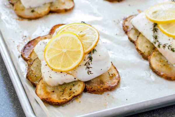 A parchment-lined baking sheet with four roasted cod fillet on top of roasted potatoes slices and topped with lemon slices.