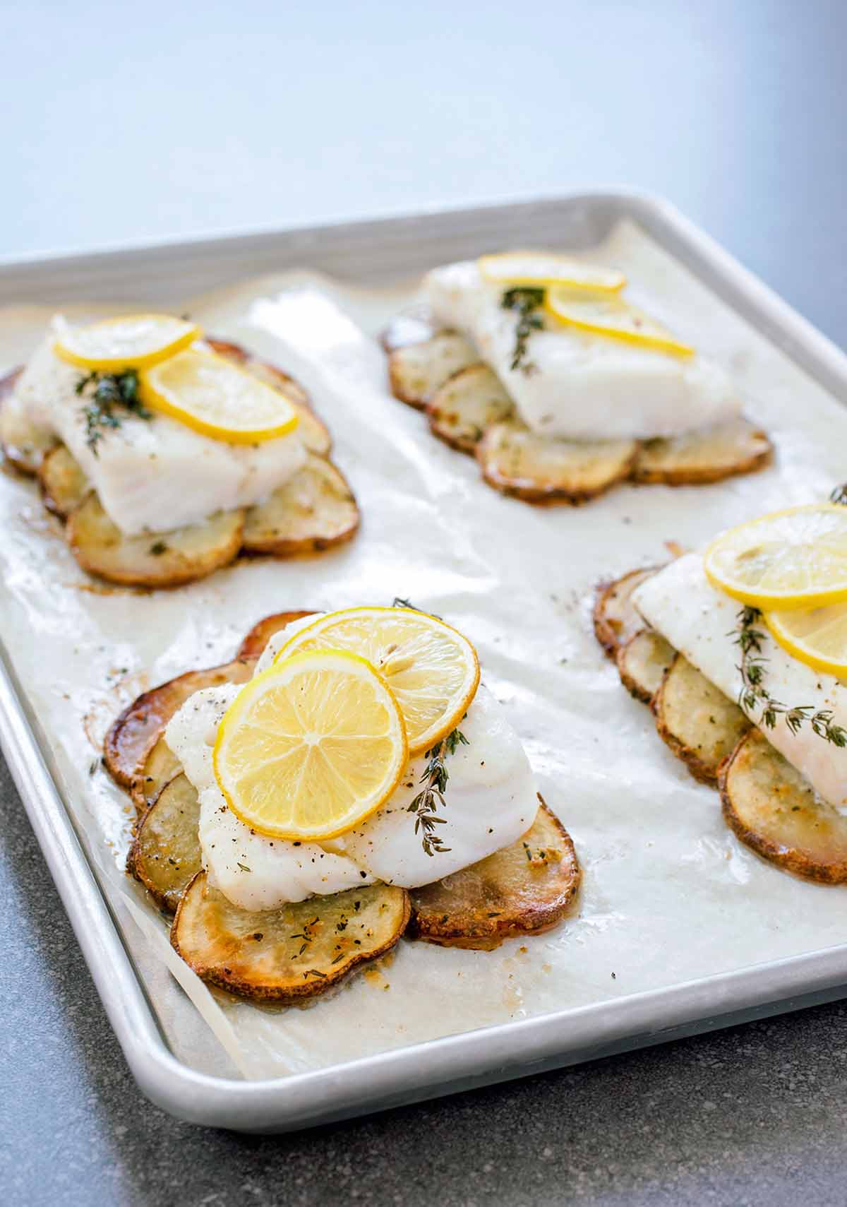 A parchment-lined baking sheet with four roasted cod fillet on top of roasted potatoes slices and topped with lemon slices.