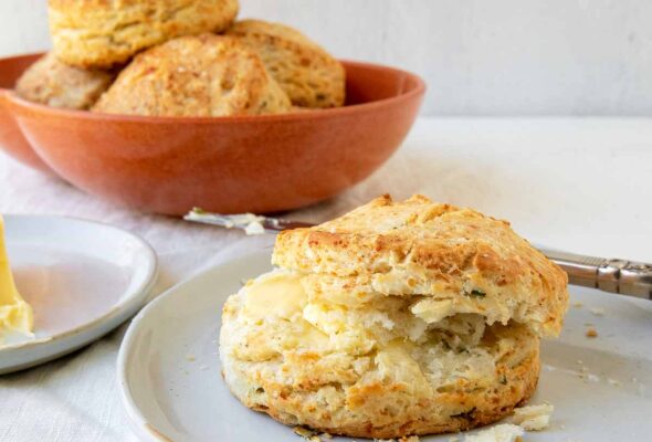 A savory Parmesan cheese biscuit with butter on a plate and a bowl of biscuits in the background.