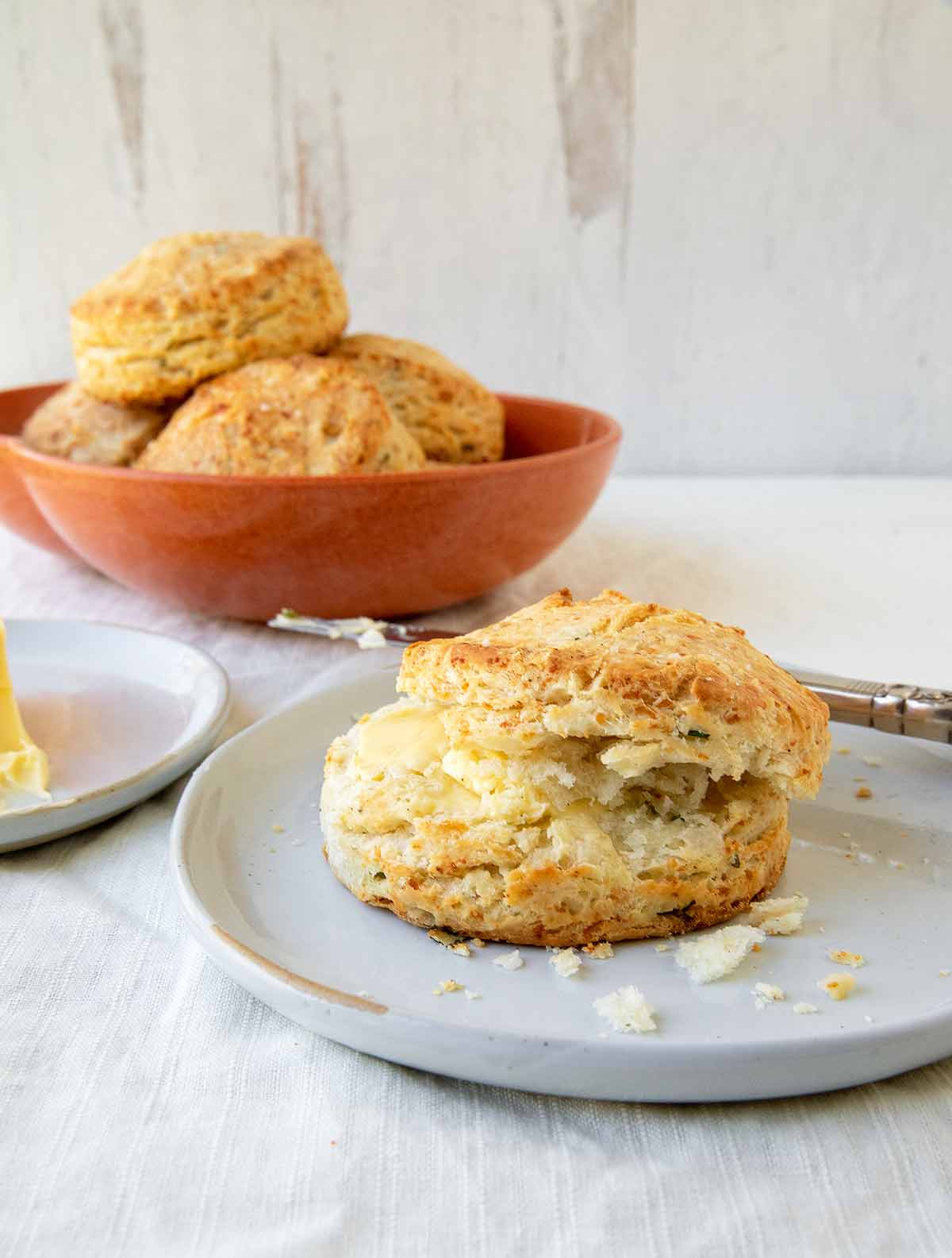 A savory Parmesan cheese biscuit with butter on a plate and a bowl of biscuits in the background.