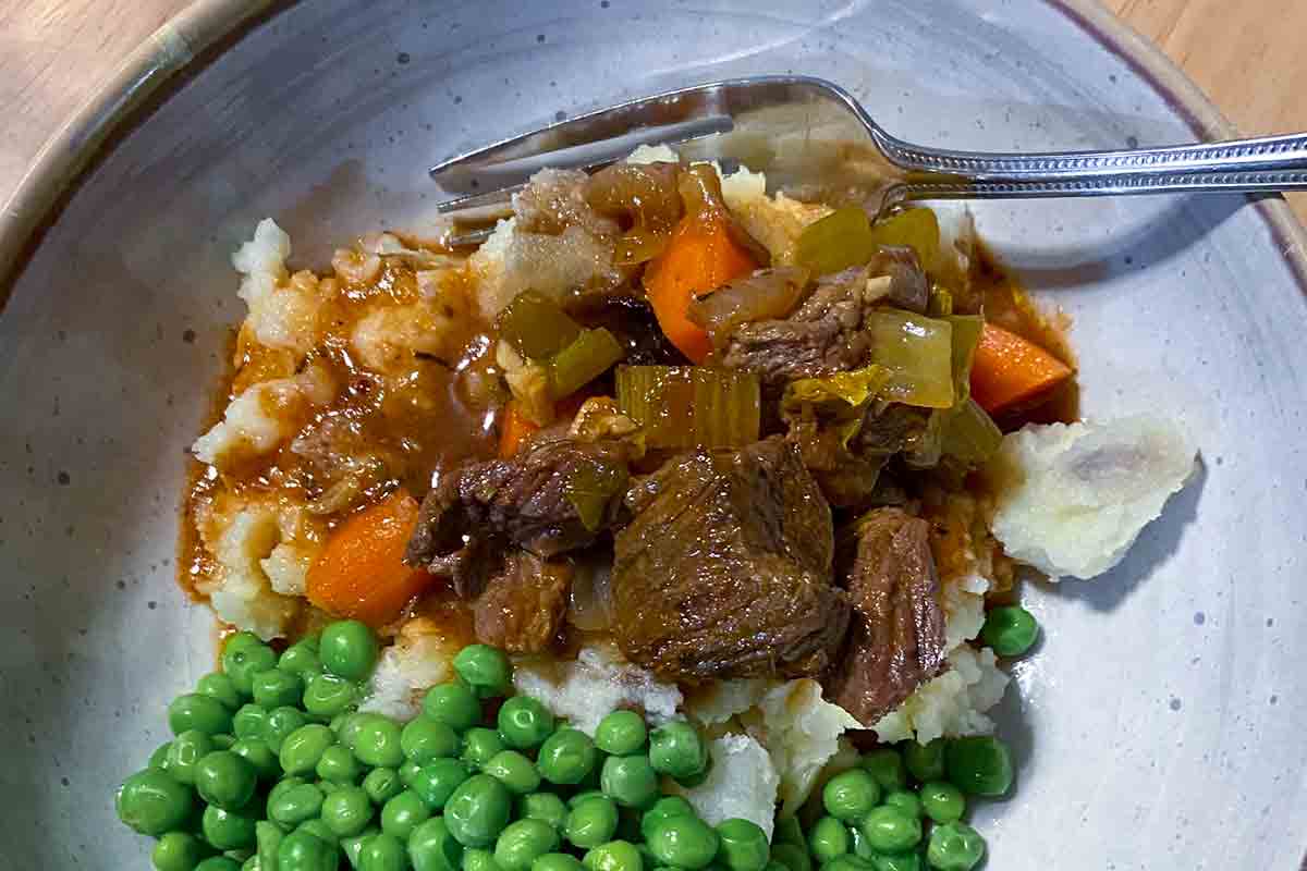 A bowl filled with mashed potatoes topped with beef stew and peas on the side.