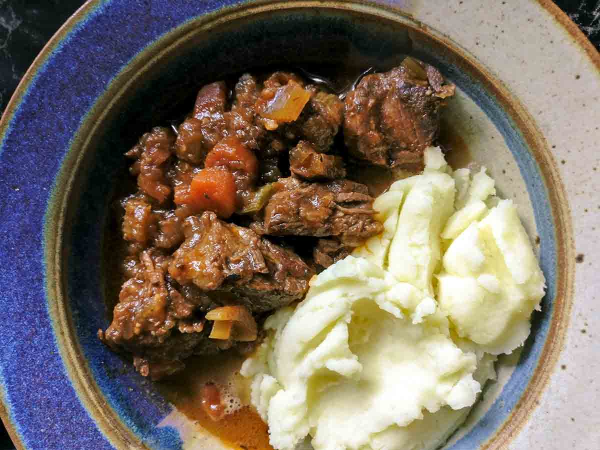A bowl half filled with beef stew and half filled with mashed potatoes.