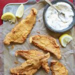Several pieces of Texas fried catfish, a bowl of tartar sauce, and lemon wedges on a metal tray lined with parchment.