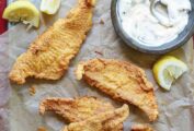 Several pieces of Texas fried catfish, a bowl of tartar sauce, and lemon wedges on a metal tray lined with parchment.