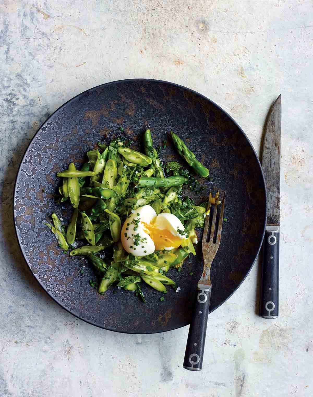 Asparagus and herb salad with chopped parsley, basil, tarragon, and mint and a broken soft boiled egg on a plate, fork and knife.
