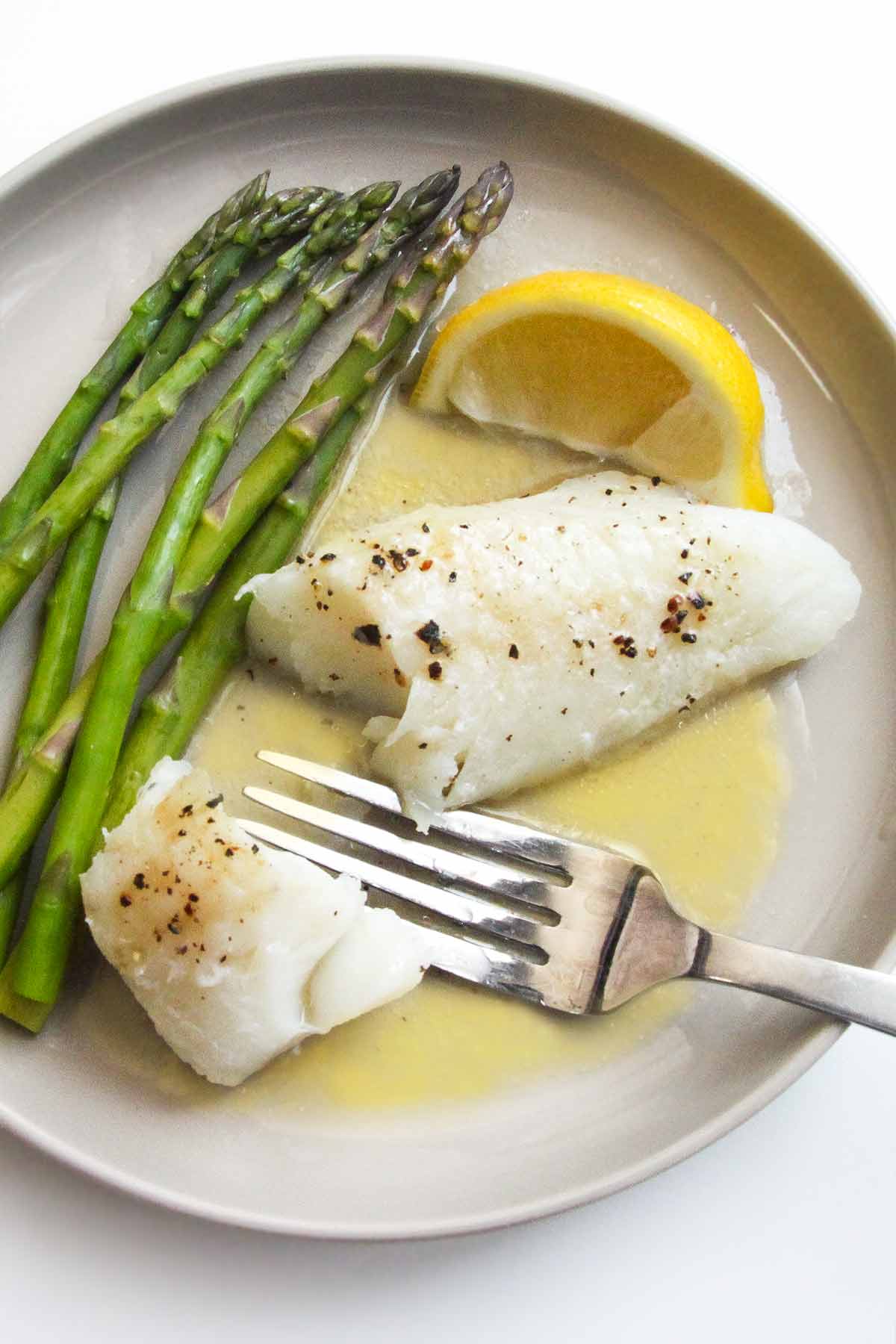 A piece of baked fish on a plate with butter sauce, a lemon wedge, and some asparagus spears.