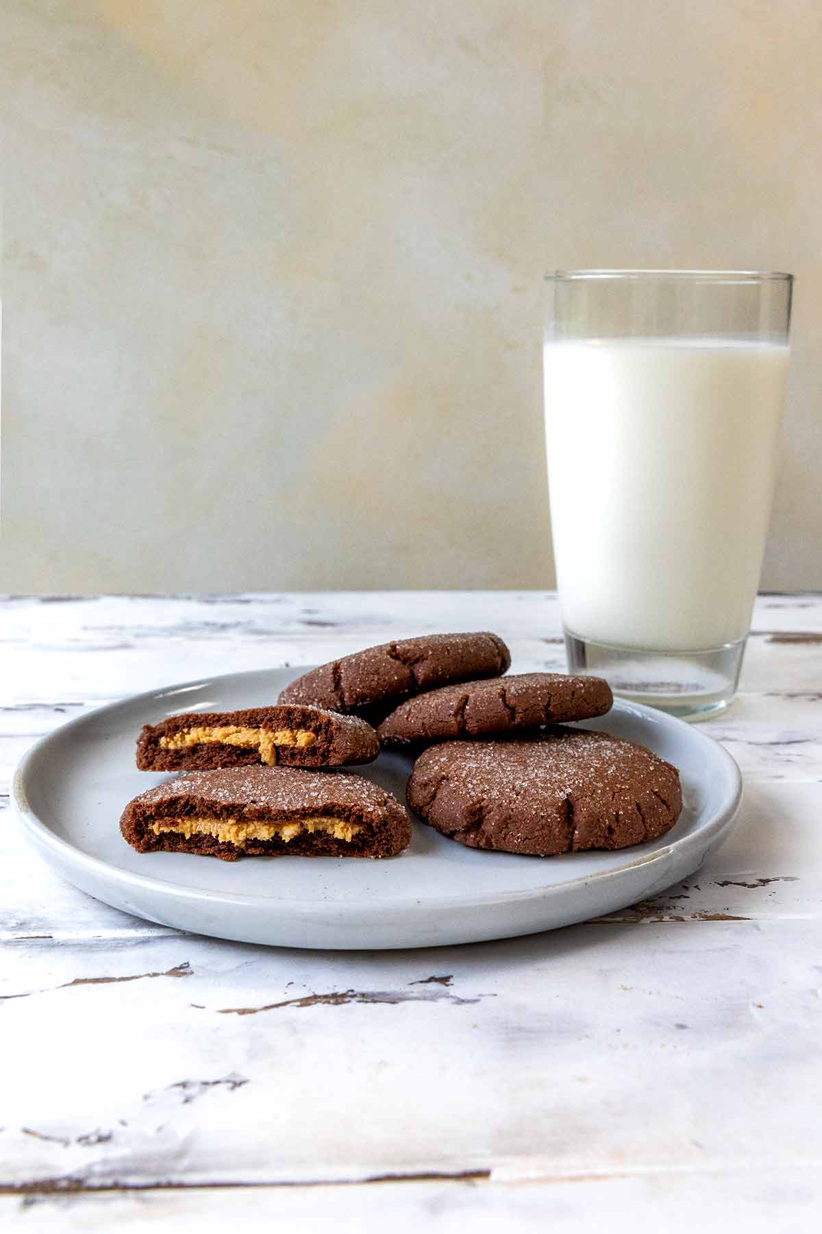 Chocolate peanut butter cookies in a stack, the top one broken in half and a glass of milk on a worn white background.