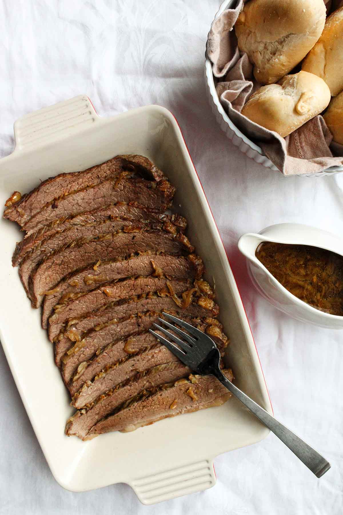 A sliced beef brisket in a baking dish with a gravy boat and basket of bread rolls on the side.