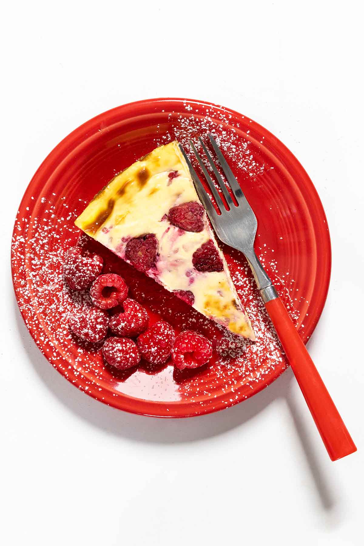 A slice of lemon raspberry cheesecake dusted with confectioners' sugar on a red plate with a fork and seven raspberries on the side.