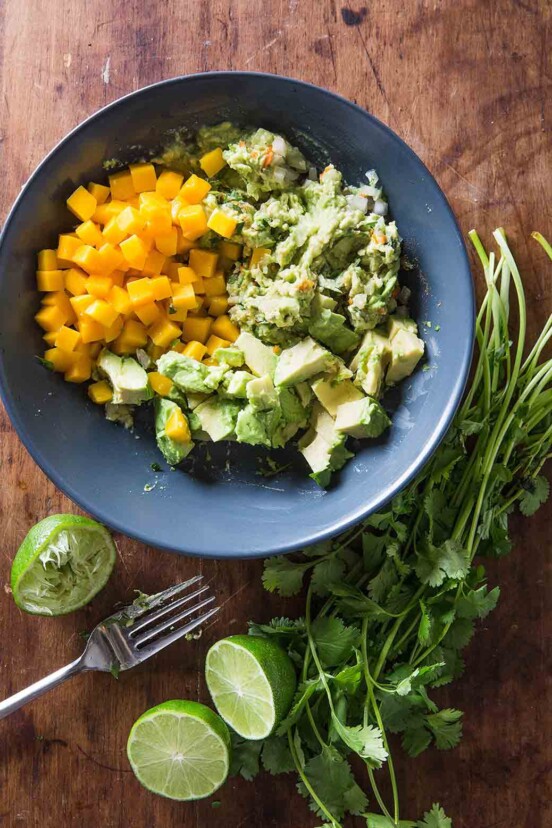 A blue bowl filled with the components of mango guacamole, with a bunch of cilantro, halved limes, and a fork on the side.