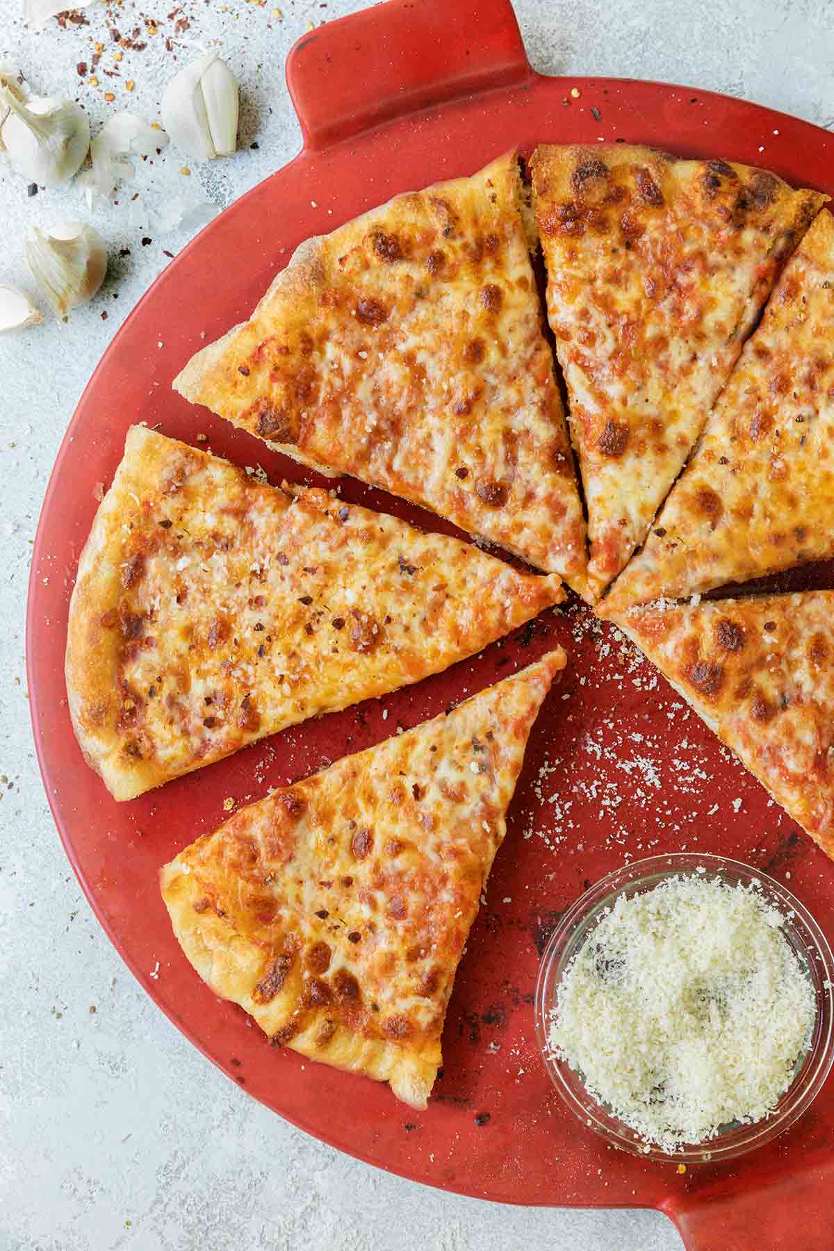 A New York style pizza cut into 8 slices with one slice missing and a bowl of Parmesan cheese on a red pizza plate.