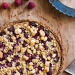 A cooked raspberry crumble tart on a wooden surface with a few raspberries lying nearby.