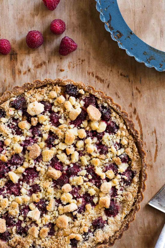 A cooked raspberry crumble tart on a wooden surface with a few raspberries lying nearby.