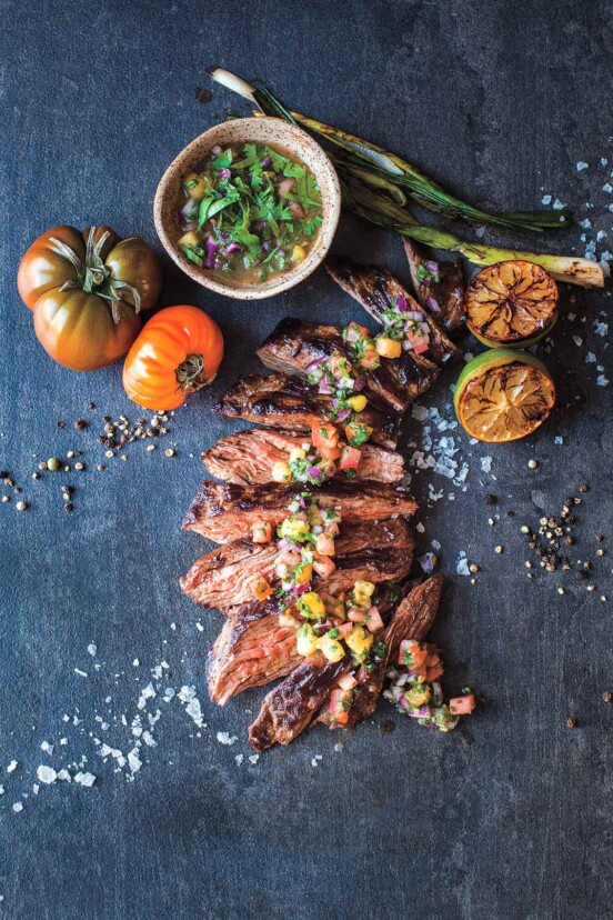 A sliced seared skirt steak with salsa dribbled over the top and a bowl of salsa, grilled limes and scallions, and tomatoes nearby.