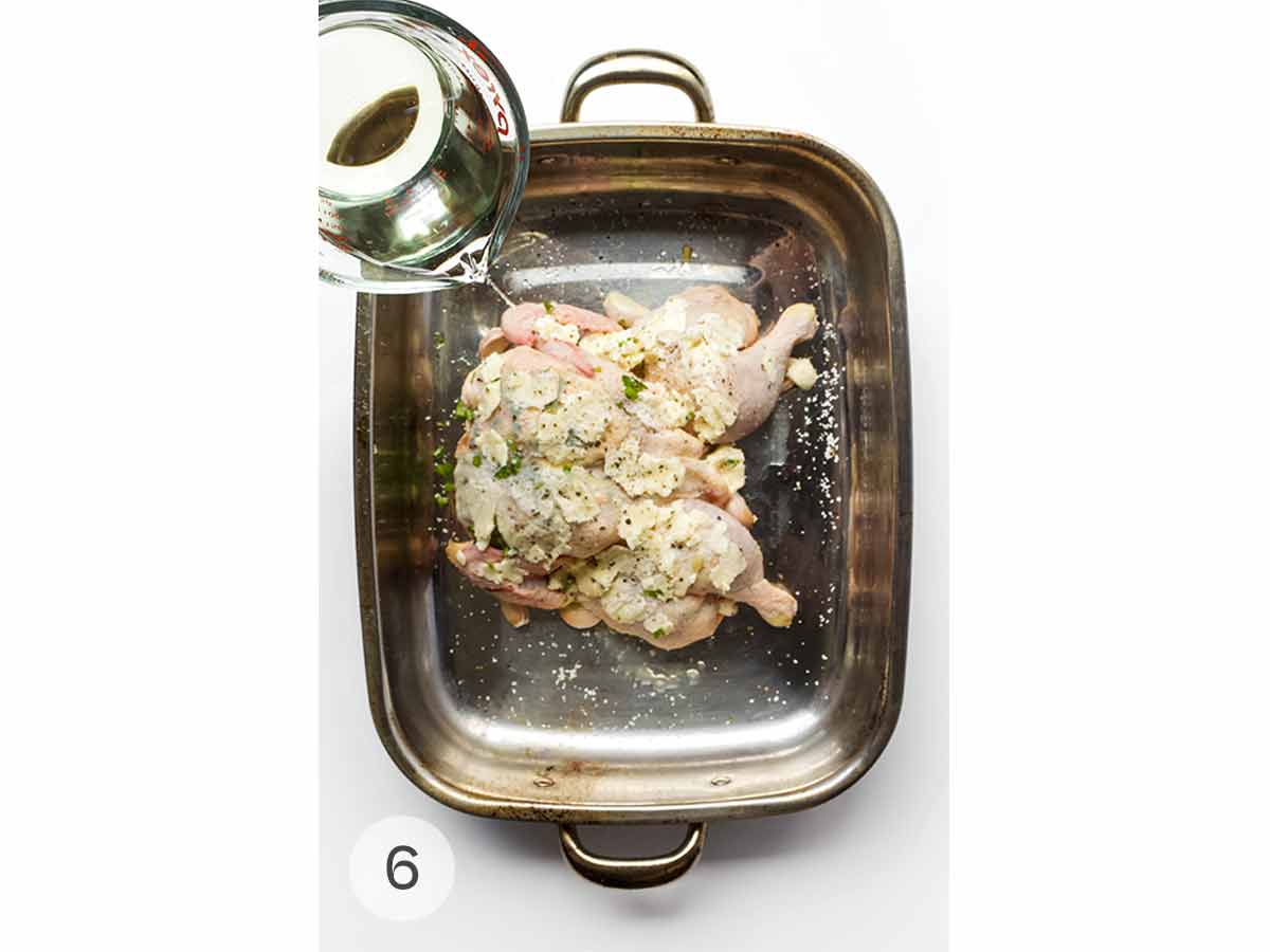 White wine being poured into a roasting pan with a spatchcocked chicken covered in butter in it.