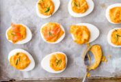 Sixteen Sriracha deviled egg halves and a teaspoon on a parchment-lined baking sheet.