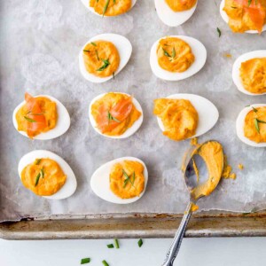 Sixteen Sriracha deviled egg halves and a teaspoon on a parchment-lined baking sheet.