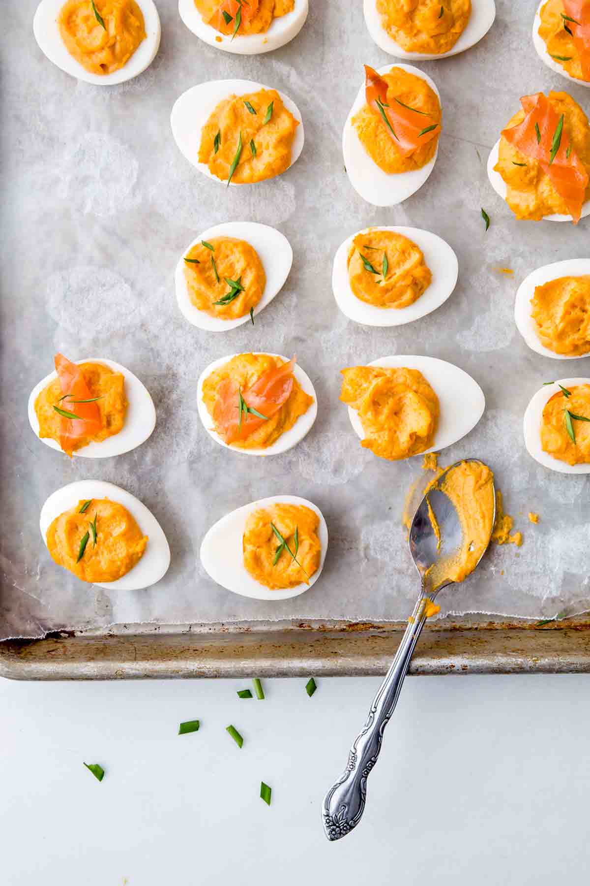 Sixteen Sriracha deviled egg halves and a teaspoon on a parchment-lined baking sheet.