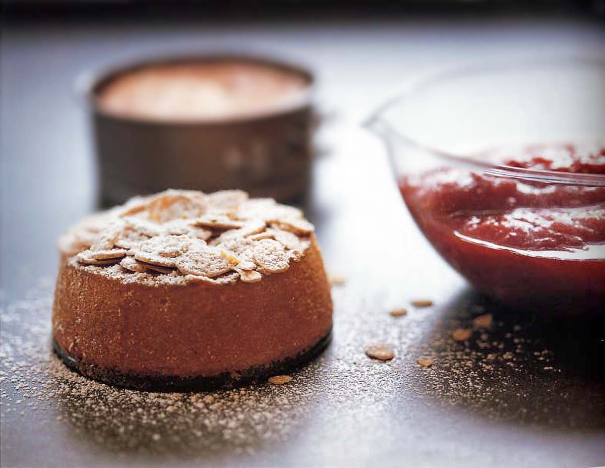 Mini almond cake topped with sliced almonds and powder sugar, nearby a bowl of strawberry-rhubarb sauce.