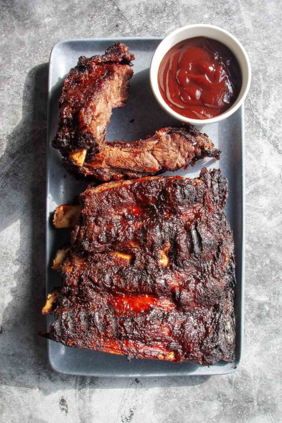 A rack of barbecued beef back ribs on a rectangular platter with a dish of barbecue sauce on the side.