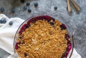 A blueberry crumble in a glass pie dish with a basked of blueberries and several spoons next to it.