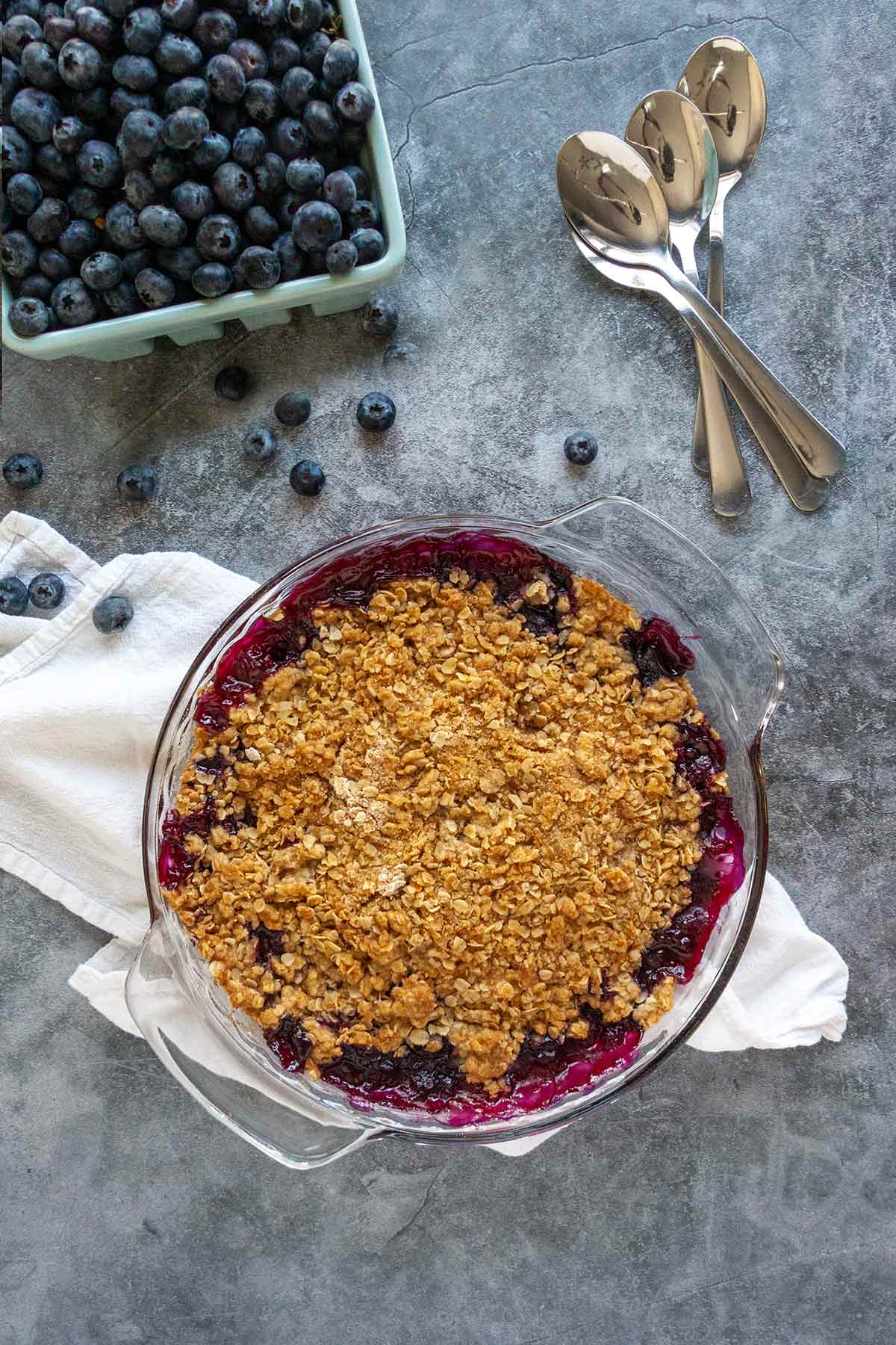 A blueberry crumble in a glass pie dish with a basked of blueberries and several spoons next to it.