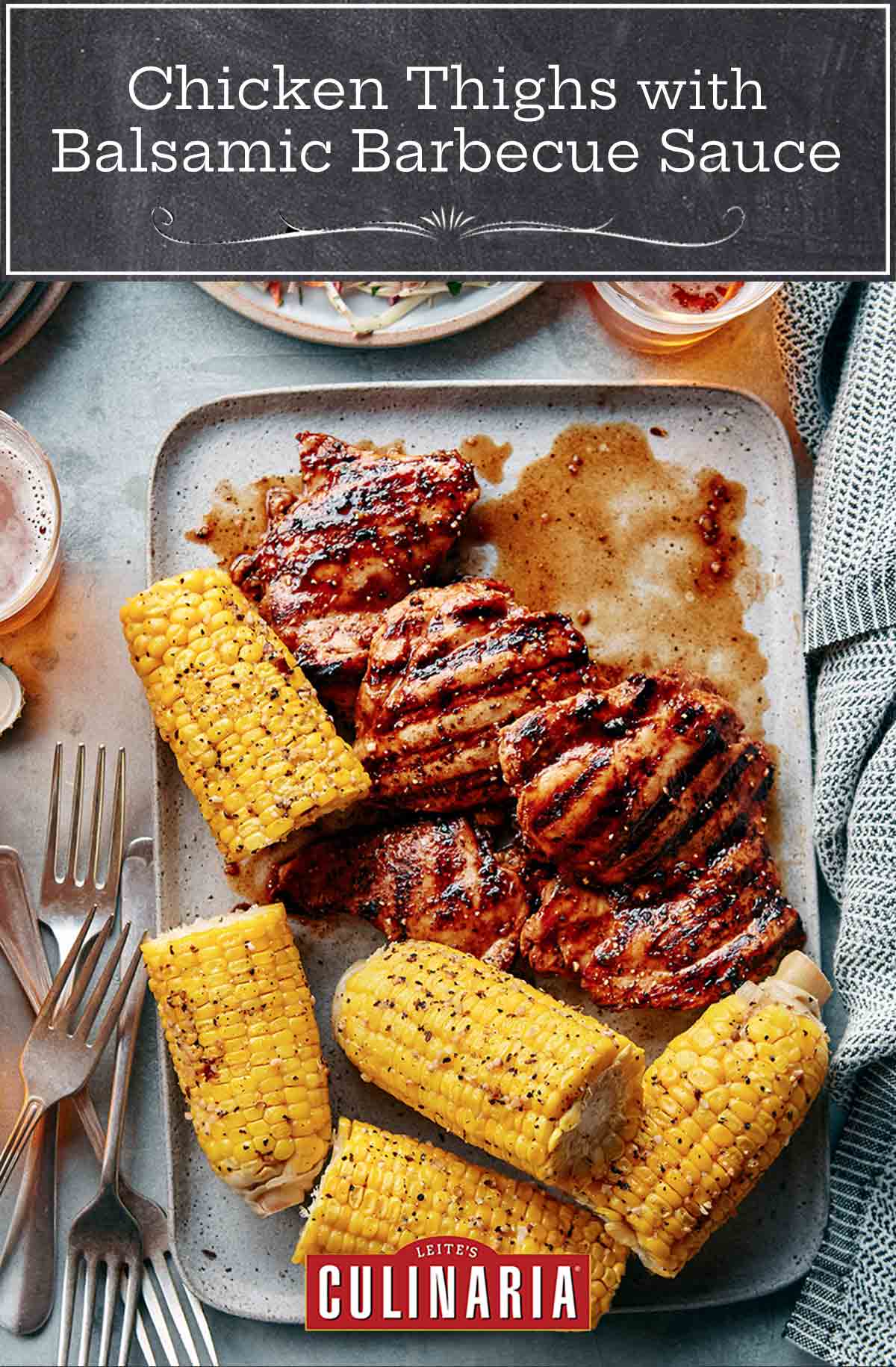 A tray of chicken thighs with balsamic barbecue sauce, corn on the cob, glasses of beer, forks, and plates.