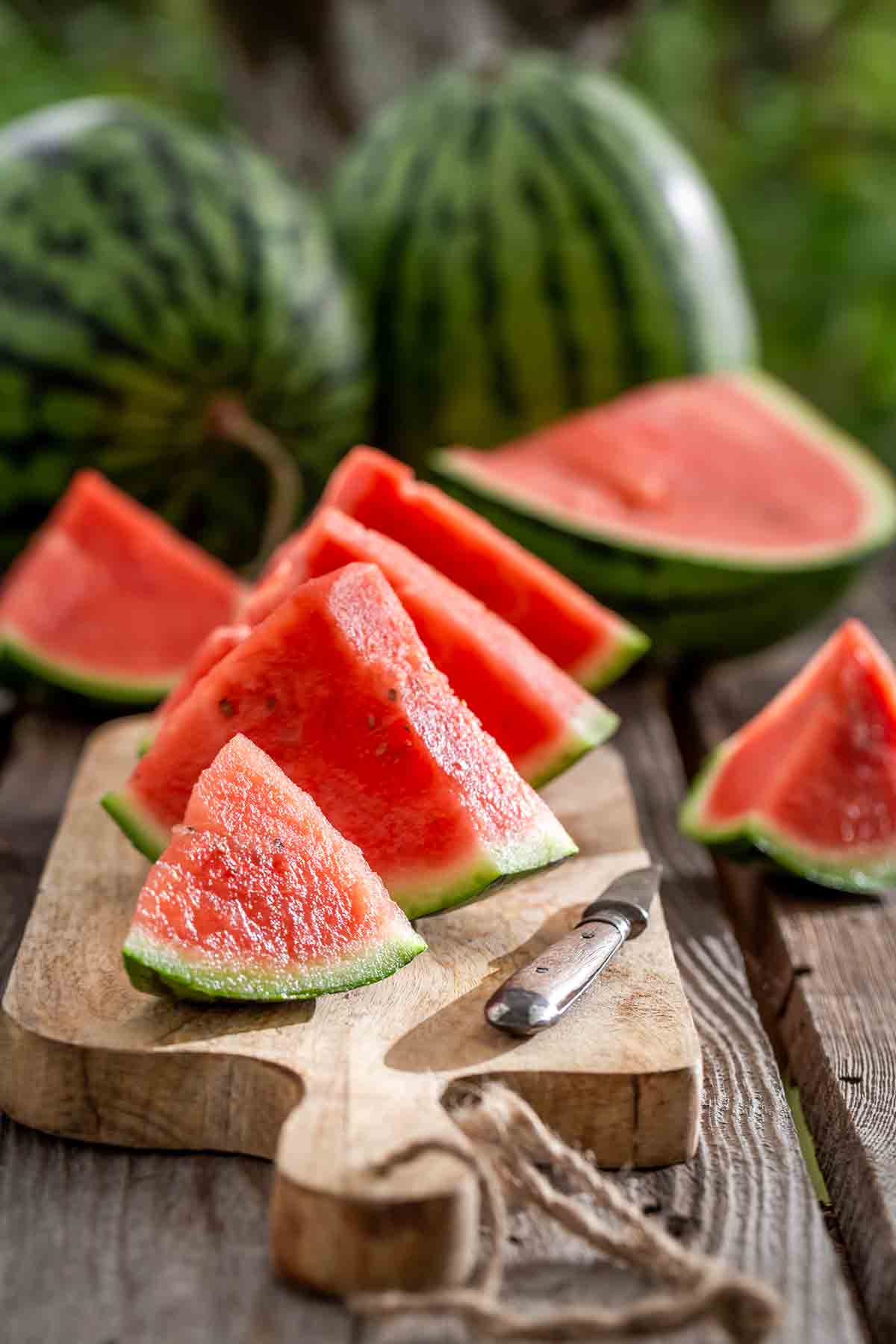 Wedges of watermelon on a cutting board, a knife on the side, and watermelons in the background.