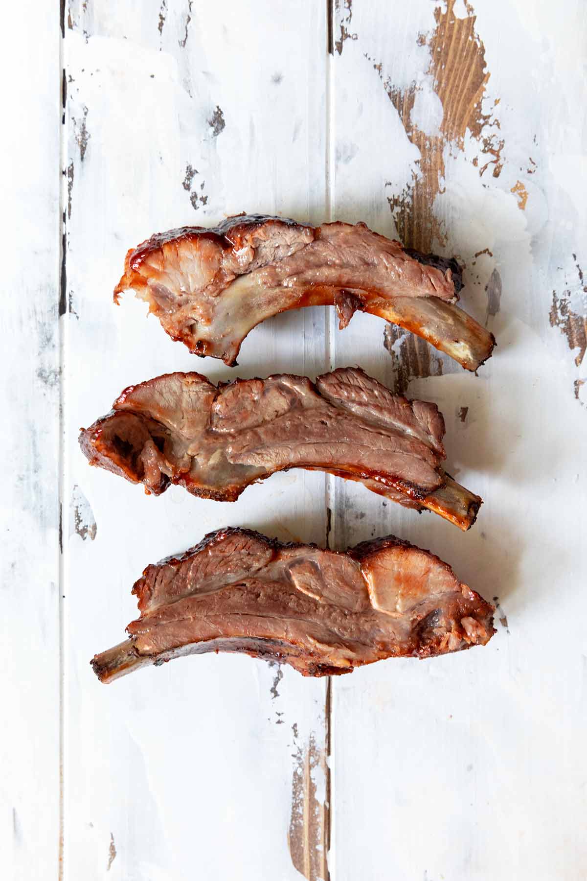 Three baby back ribs on a worn white wooden table.
