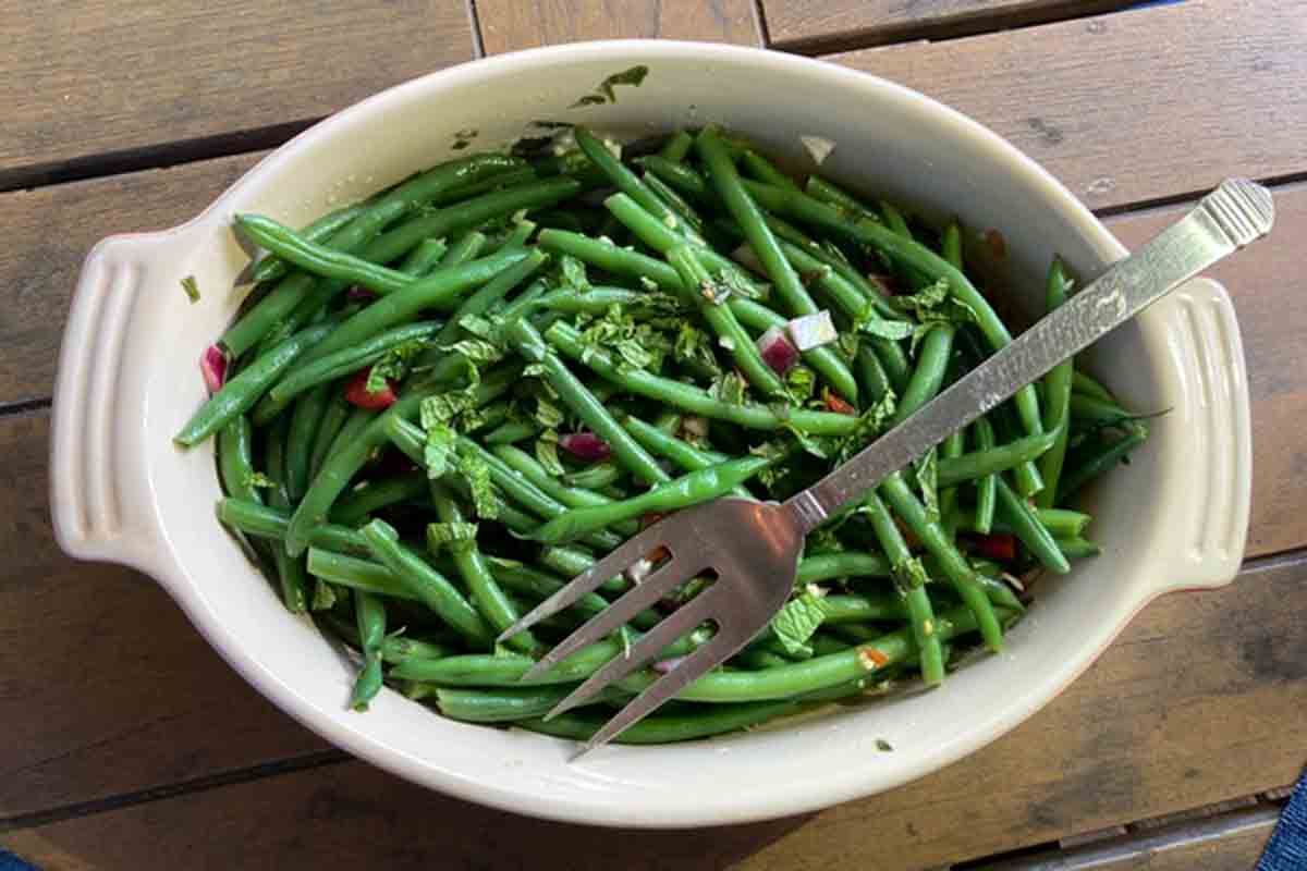 An oval dish filled with green bean salad, red onion, and fresh mint, with a fork resting on top.