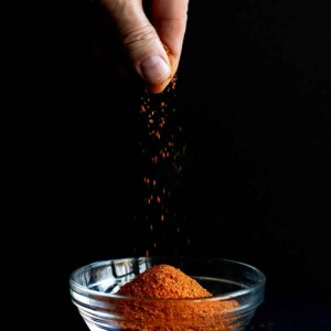 A hand sprinkling homemade Old Bay Seasoning in a small glass bowl.