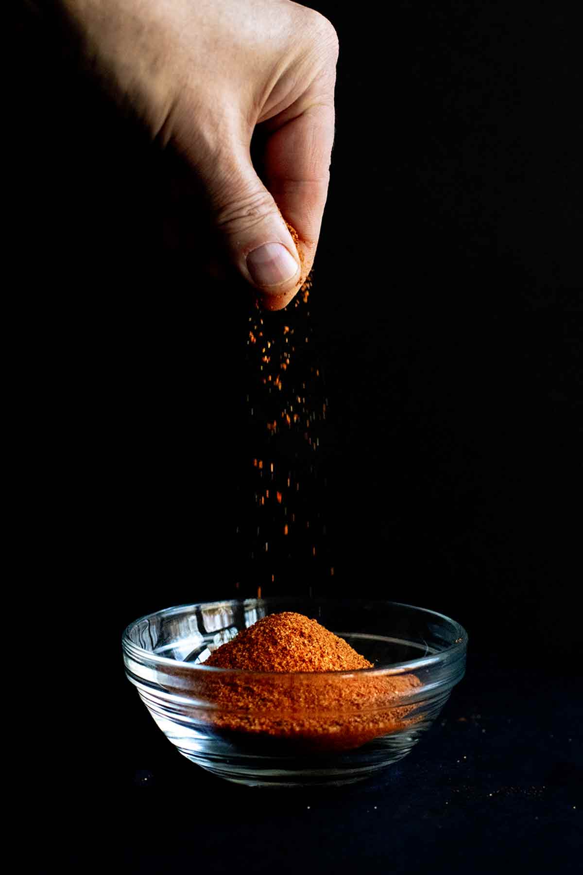 A hand sprinkling homemade Old Bay Seasoning in a small glass bowl.