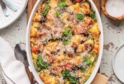 A breakfast casserole in an oval white staub dish on a wooden board with plates, Parmesan, cilantro, and a dish of salt on the side.