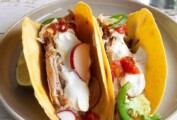 Two pork tacos filled with shredded pork, radishes, jalapenos, salsa, and sour cream on a plate.