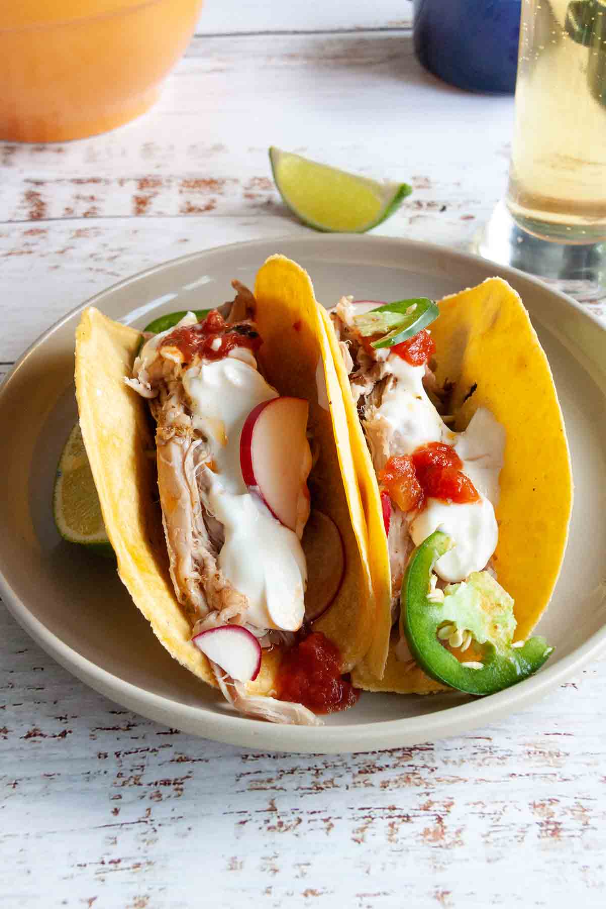 Two pork tacos filled with shredded pork, radishes, jalapenos, salsa, and sour cream on a plate.