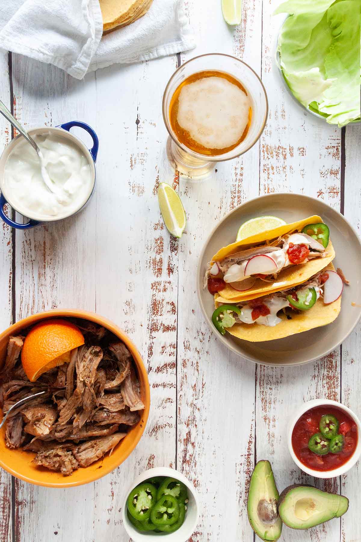 Zwei Schweinefleisch-Tacos, eine Schüssel mit zerkleinertem Schweinefleisch und eine Auswahl an Gewürzen auf einem weißen Holztisch.