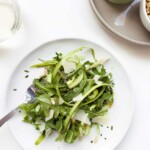 A plate of raw asparagus salad with a jar of dressing and a bowl of pine nuts, and a glass of white wine on the side.