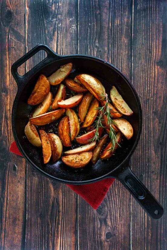 A cast-iron skillet filled with roasted grilled potatoes and two sprigs of rosemary on a wooden table.